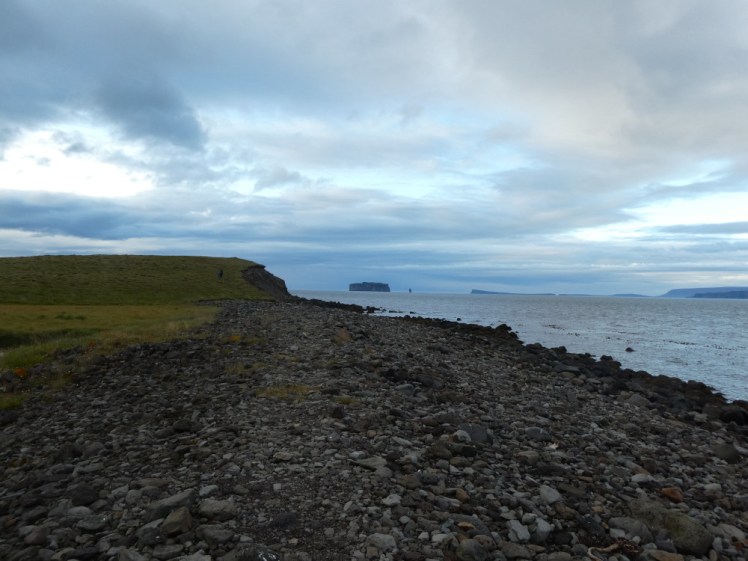 A bit of black rocky shoreline rising up into a green hill at the end. Far away in the distance is a square-sided island, some four or five miles out to sea, which Grettir once swam from.