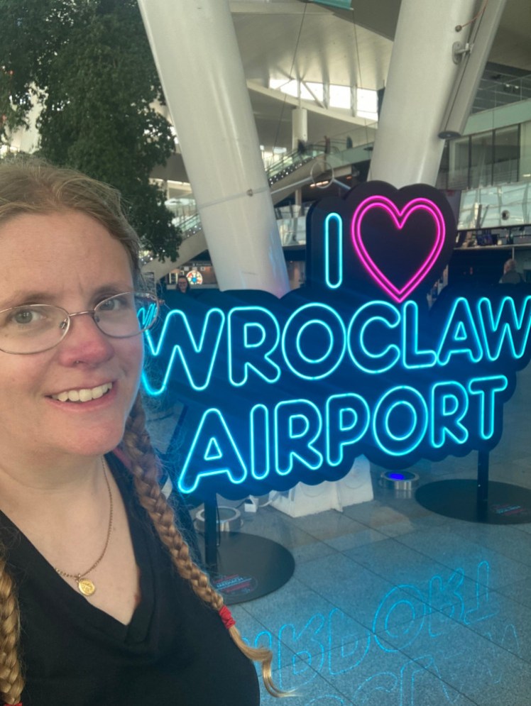 A selfie looking sceptical next to a neo sign that says I heart Wroclaw Airport.