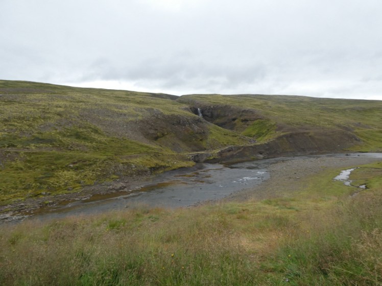 A small waterfall in a hollow in the hillside. A shallow river wends its way along the foot of the hillside and joining the waterfall and river is a concrete bridge, so small and old-fashioned that you can hardly see it. That was once the main road to Akureyri.