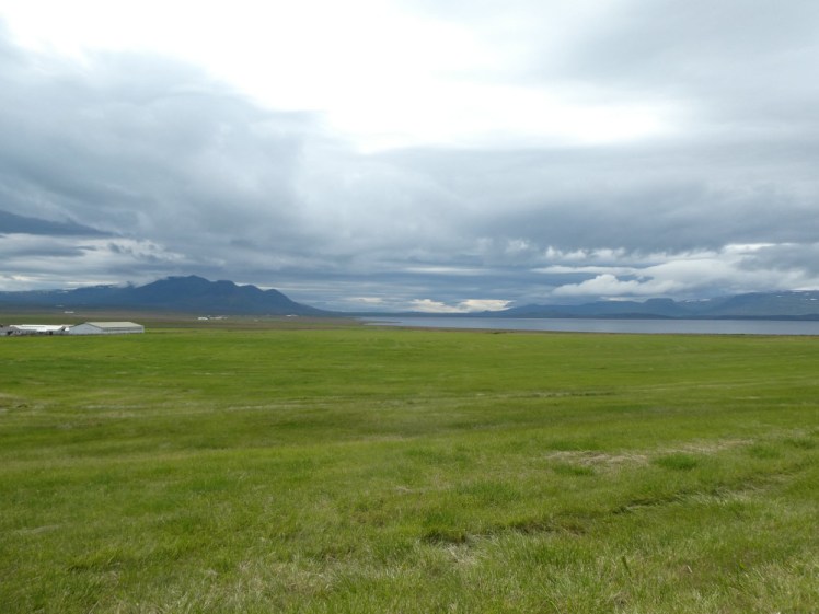 An expanse of grass with a fjord gleaming in the distance, a mountain rising up on the left and heavy black clouds looming over it all.