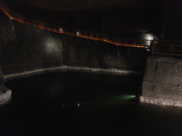 A salt lake in deep green, lightly lit around the edge. Above it, a walkway lit in orange goes up the wall.