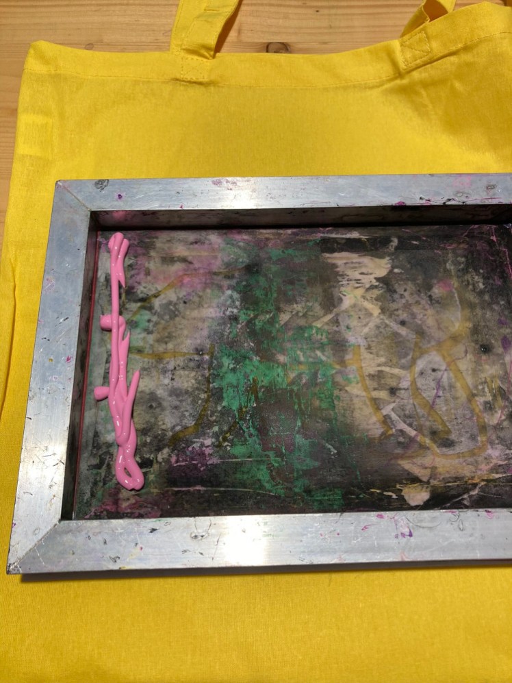 Screenprinting - a screen laid down on top of a yellow cotton bag with a squirt of bright pink paint on it.