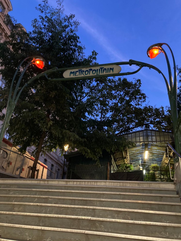 Emerging at Cité station while the sky is still royal blue and the orange lights over the entrance are still orange. It has the traditional Parisian Art Deco Metropolitain sign over the stairs.