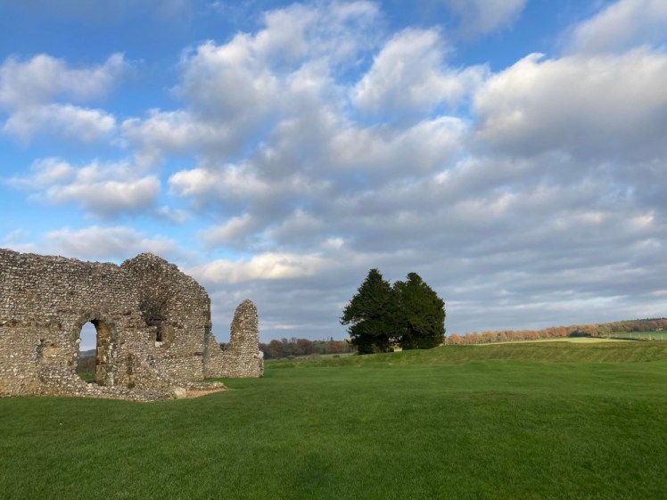 A ruined church wall and the ramparts behind it. Beyond the ramparts is a clump of trees, which are within a large barrow, although you can't see it because of Church Henge's ramparts.