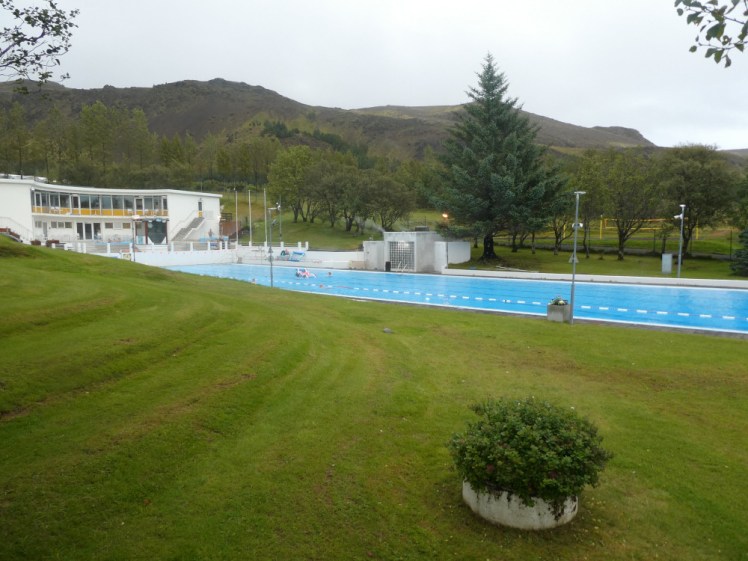 A long blue swimming pool set in something that looks like lawn. At the far end, a two-storey curved building hugs the small pool and hot tub, neither of which you can see from this angle.