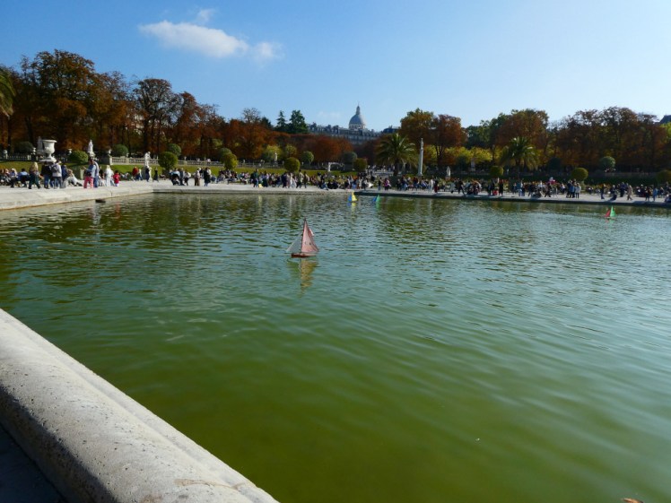 The fountain of the Jardin du Luxembourg with toy boats sailing around in it. Behind it is lots of greenery.