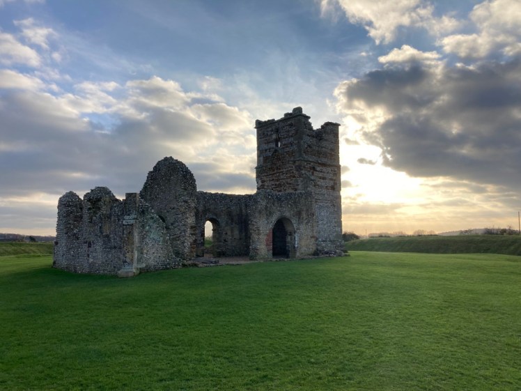 Knowlton Church, a ruin of a medieval church in a field of very green grass with the sun behind it, about to vanish behind a heavy grey cloud.