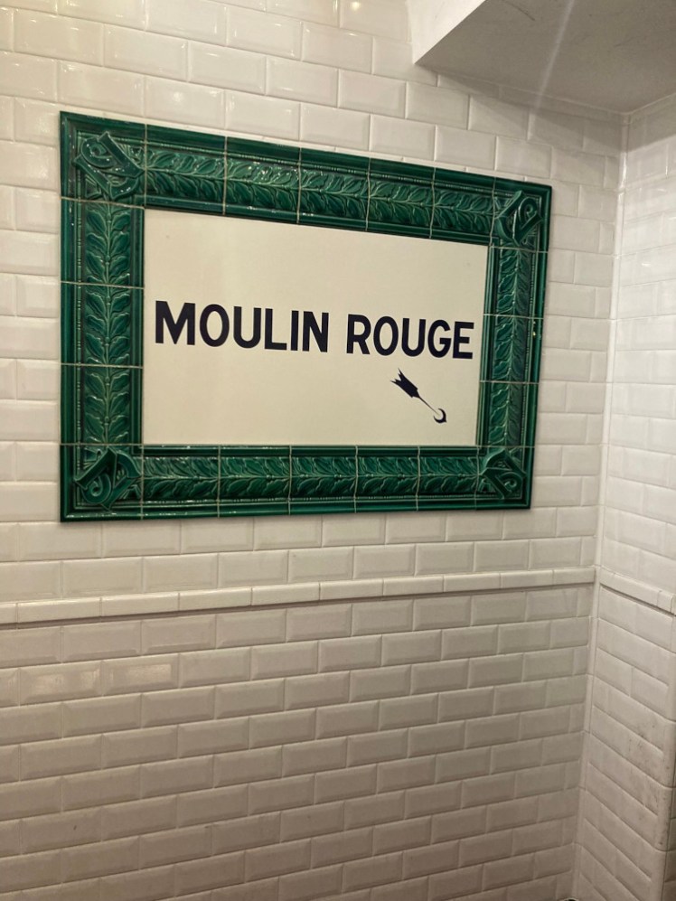 A Moulin Rouge sign in tiles with a green border on a white brick background. The effect is of sneaking into some hidden secret underground club.