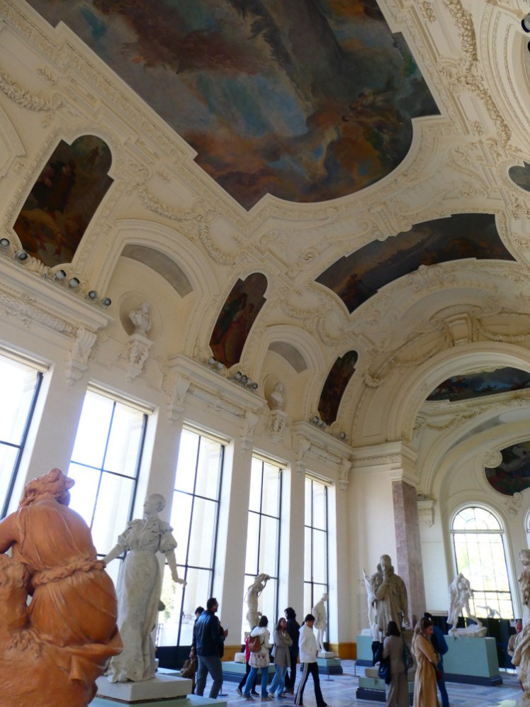 Inside the Petit Palais. The ceiling is painted with large oils and the floor is littered with small sculptures and people walking among them.
