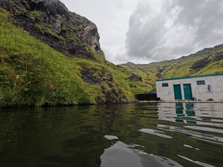 A slightly murky dark green pool with a shabby off-white building at the end. From this angle, the lush green mountains rise directly out of the pool.