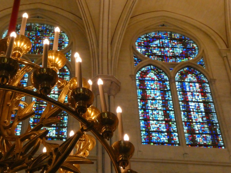 A stained glass window high up, made up of lots of little squares (possibly from the scraps of windows exploded in the fire) and a multi-level gold chandelier.