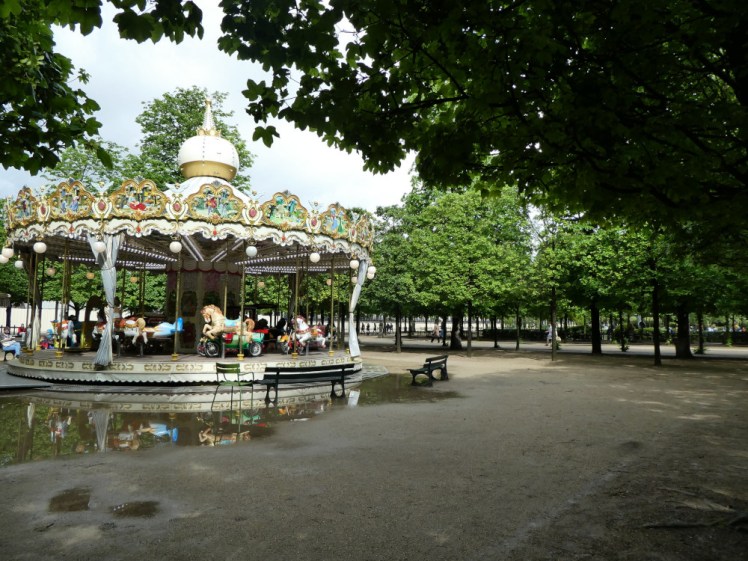 A carousel in the Tuileries Gardens. The trees are vivid green and the ground is very wet from recent rain.