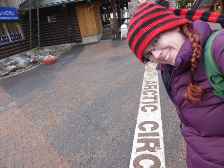 A selfie at the Arctic Circle, depicted by a white line painted on the ground. I'm wearing a purple padded coat, a long-tailed red and black striped coat and my hair dyed red in plaits falling over the purple coat in a really mismatched way.