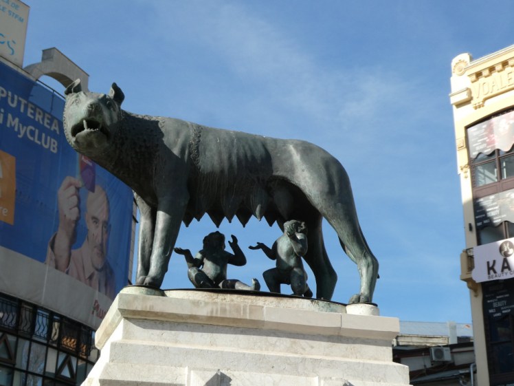 A bronze statue of a female wolf feeding two human babies, Romulus and Remus, founders of Rome. This version, in Bucharest, is in a fairly busy square with shops & adverts visible behind it.