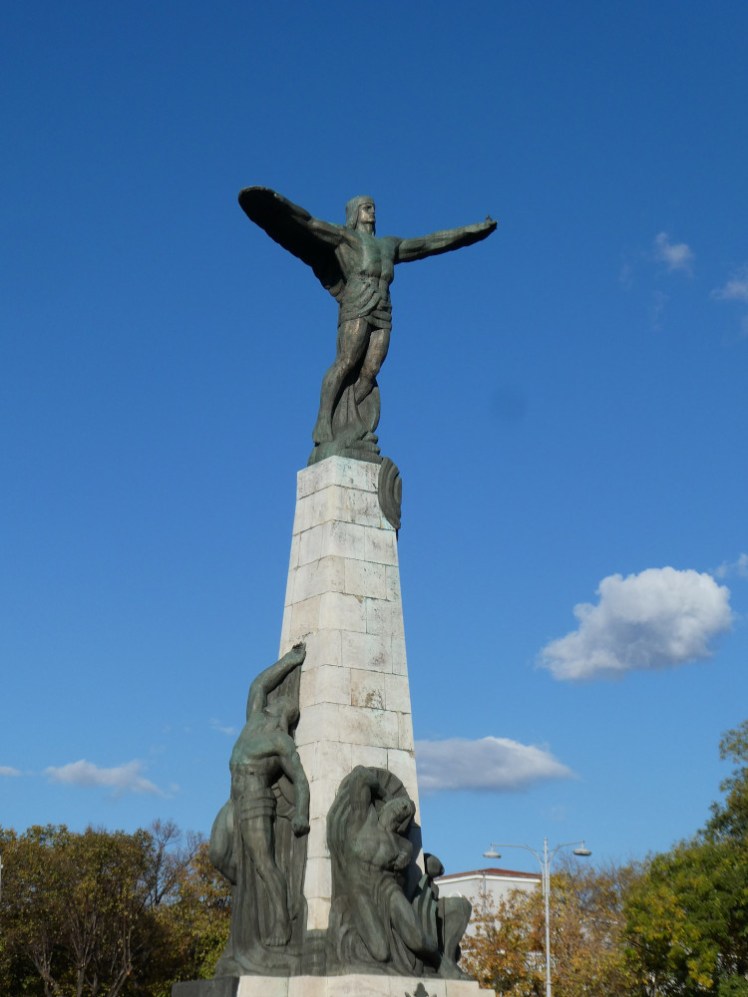A stone column topped by a man with his arms outstretched and a shawl hanging from them, which mostly just make him look like he has very wide arms or very small wings.