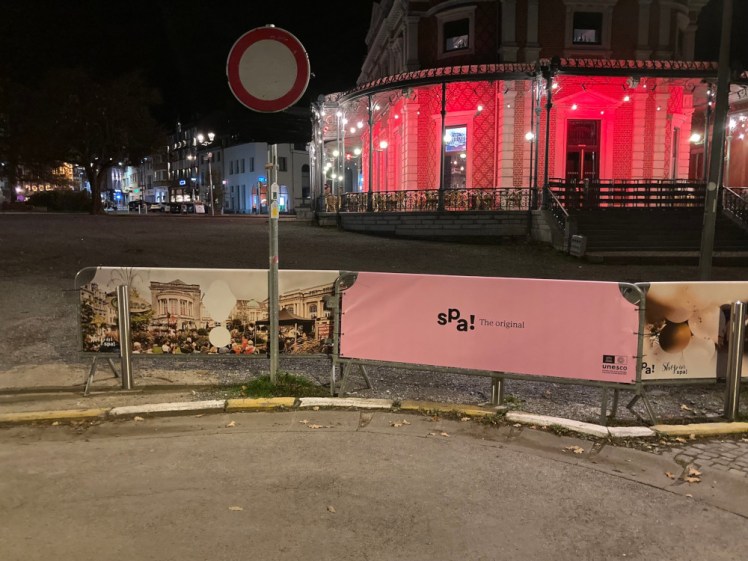 Some temporary fencing around what will become a market. All piece of fence visible have a banner over them; the central one is light pink with Spa! on it in a jaunty font, declaring it "the original".