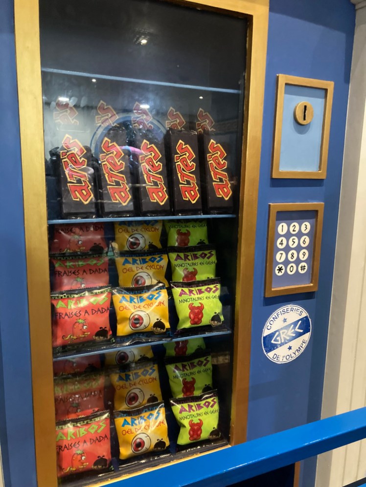 An Ancient Greek vending machine featuring bags of "Aribos" in pointed Greek-style lettering and Ares chocolate bars.