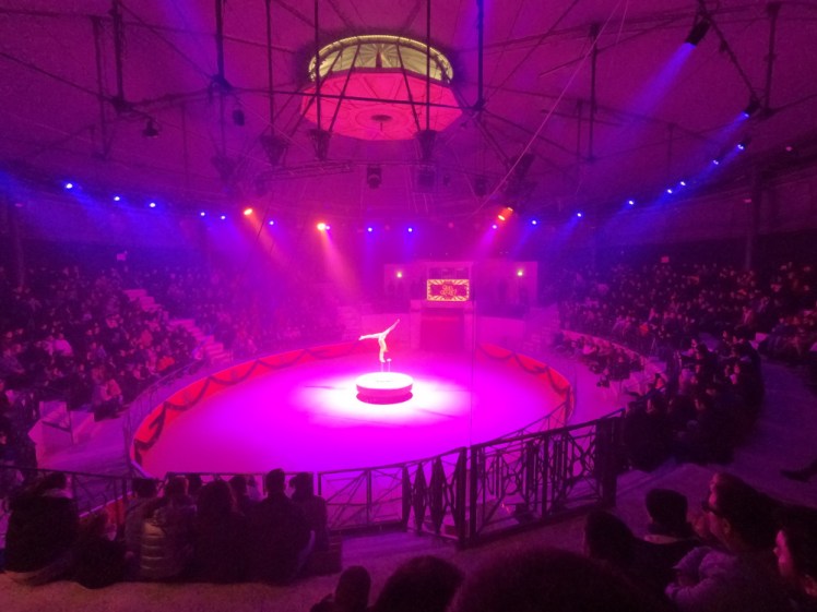 A balancing act, a man balanced on his hands on two tall thin stands as the central circular part of the ring floor rises up.