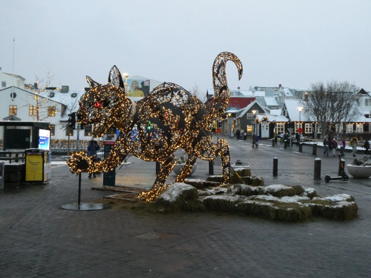 The Christmas Cat sculpture, a massive illuminated cat in central Reykjavik. Behind it, it's snowing and the roofs are covered in snow but the ground is completely clean and dry.