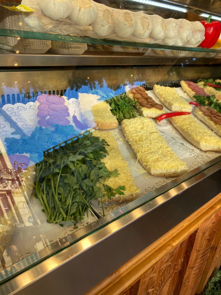 Slabs of bread covered with a thick layer of grated cheese and some with sausage or herbs piled on too. On top of the counter is a net of large bulbs of garlic, in case you weren't clear that this is garlic cheesy bread and not plain cheesy bread.