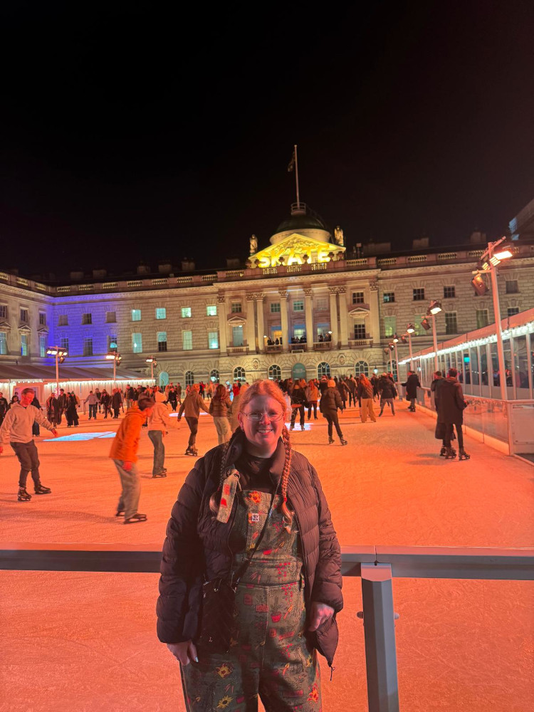 A picture of me standing leaning on the rail around the ice rink, wearing green dungarees and a down jacket looking like I have no idea how to stand like a human being when a camera is pointed at me.