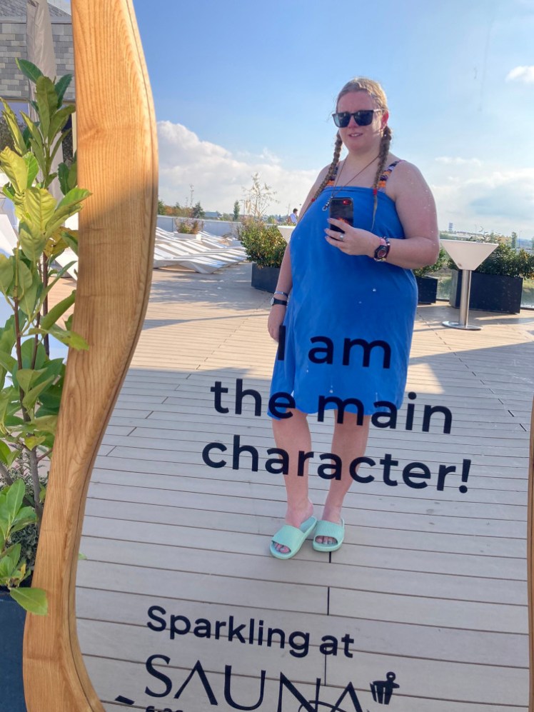 A mirror selfie wearing a blue towel and green plastic sandals. The mirror is left over from their sauna event and has "I am the main character!" written across it.