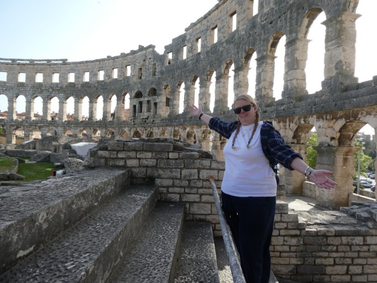 A selfie at the Roman Arena in Pula. I'm wearing a white t-shirt with a blue checked shirt and blue linen trousers holding my arms out in triumph.