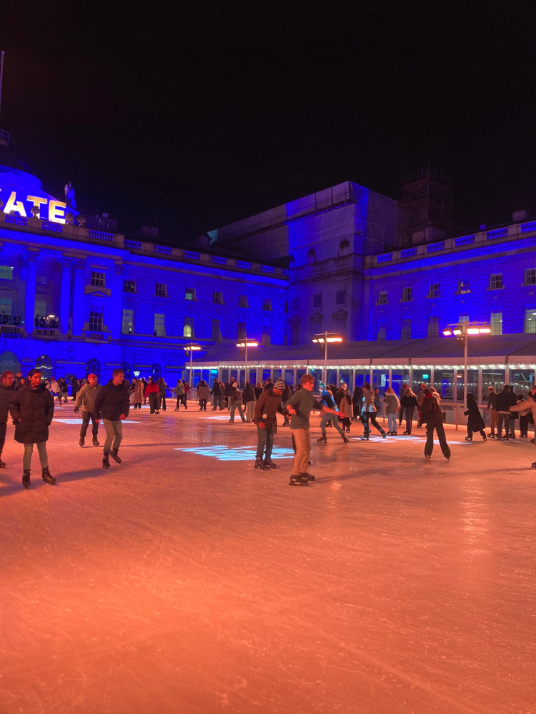 A corner of the ice rink illuminated in a kind of peach-orange light. Somerset House, behind it, is lit in bright blue.