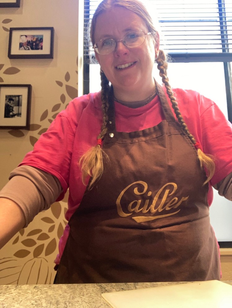 A timer selfie in my apron at my workstation. I'm wearing a bright pink t-shirt and a chocolate-brown apron with the Cailler logo embroidered on the front in gold.