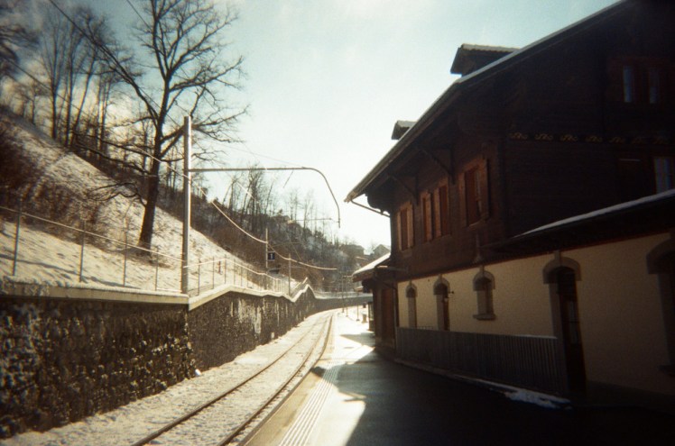 The station at Broc-Chocolaterie - a brown and white chalet housing the station building is on the right and a steep hill with a stone wall holding it back on the left. The train track runs between the two and although the house is probably darker than its digital equivalent, this isn't a bad picture either.