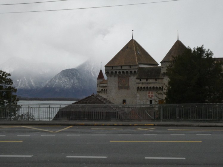 Chateau de Chillon seen from the other side of the road with misty mountains on the lake behind it.