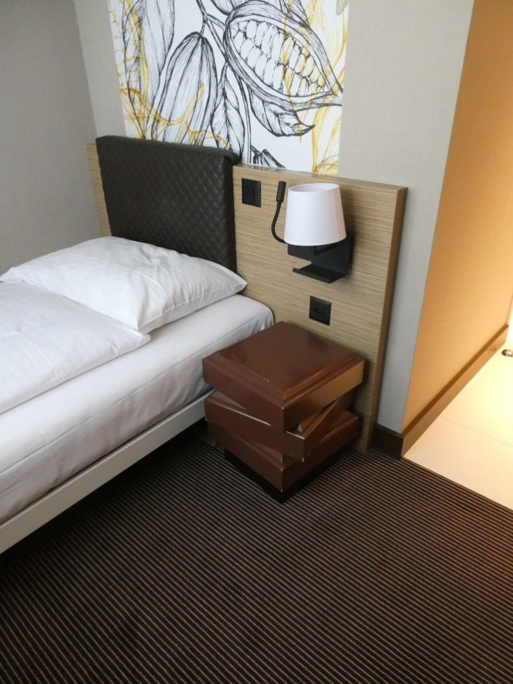 The bed, with a bedside table resembling a stack of chocolate. Behind the single bed, you can see a mural of cocoa beans which runs up the wall and across the ceiling.