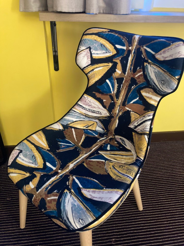 A chair upholstered in a fabric with a pattern of cocoa beans in shades of brown, yellow and blue. The wall behind it is a shocking shade of yellow.