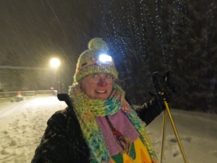 Me, slightly blurred, heading away from the station at Les Pleiades. I'm wearing my Fluffy Clouds hat and scarf with my headlight on, trying to point it away from the camera. I've got two poles in my other hand and my big black heated coat worn open over my rainbow fleece because it's too much effort to zip it up right now.