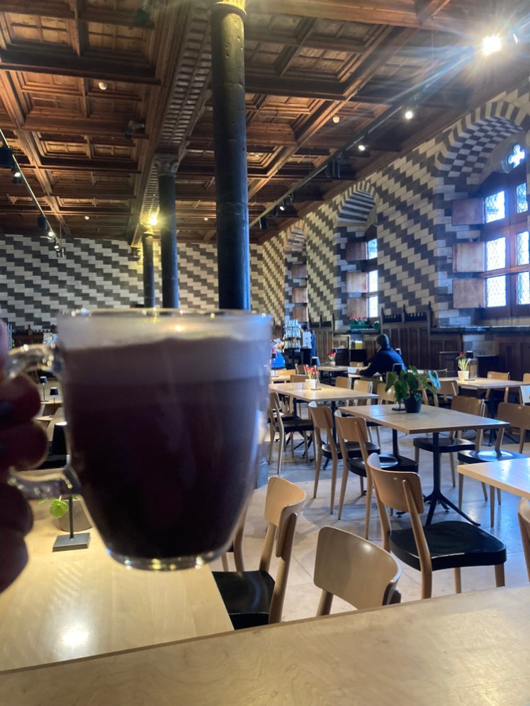 A cup of hot chocolate in a glass cup in a medieval banquet hall with stripy walls and ancient pillars.