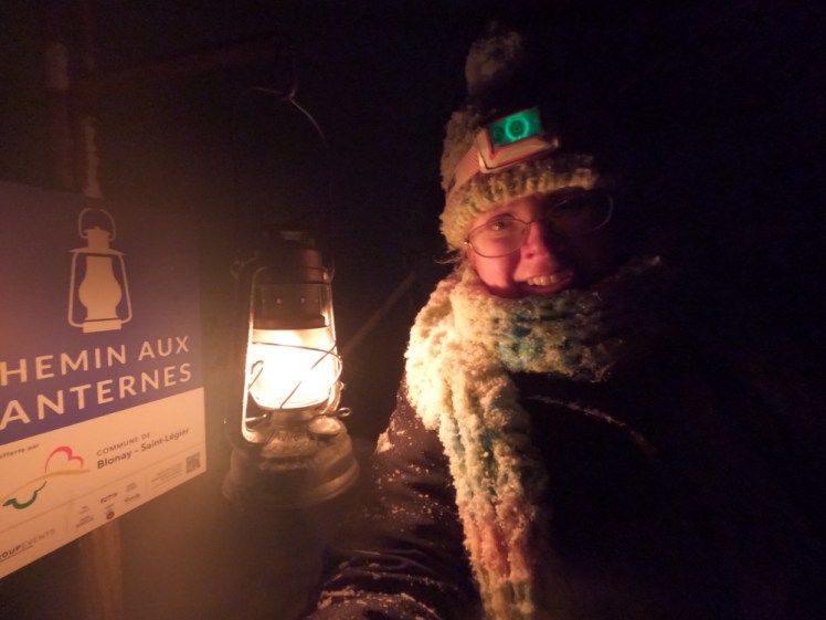 A selfie with my headtorch off, standing next to one of the lanterns and the Chemin aux Lanternes sign that explains what's going on.