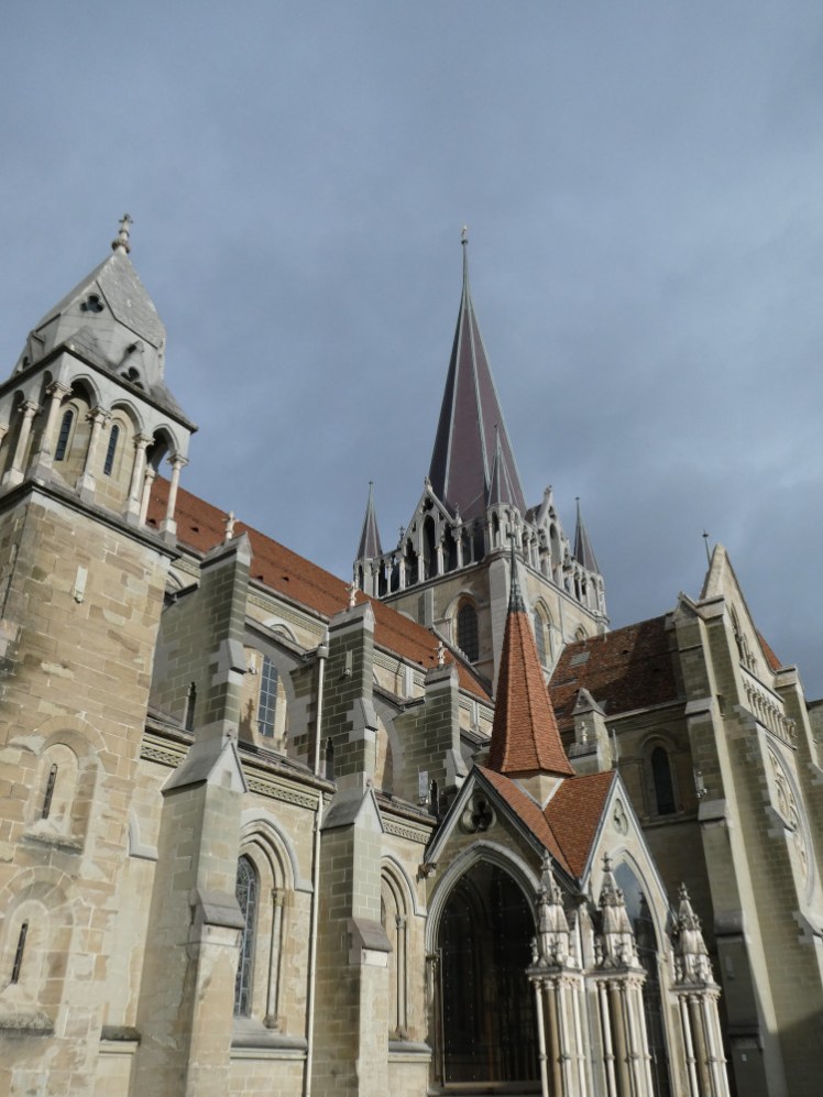 Lausanne Cathedral as seen from human eye level with a cloudy but vaguely blue sky above it. It's early Gothic, with decorations on the tower and dark orange roof tiles on all its slanted roofs. The stone itself is a slightly yellow local sandstone.