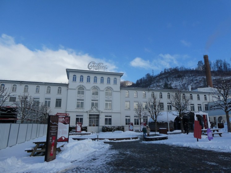 Maison Cailler, a factory that looks more like a classical-style mansion. The centrepiece is a four story white building with decorative moulding around the big windows and then there are three-storey wings sticking out on both sides. The whole is crowned with the Cailler word-logo.