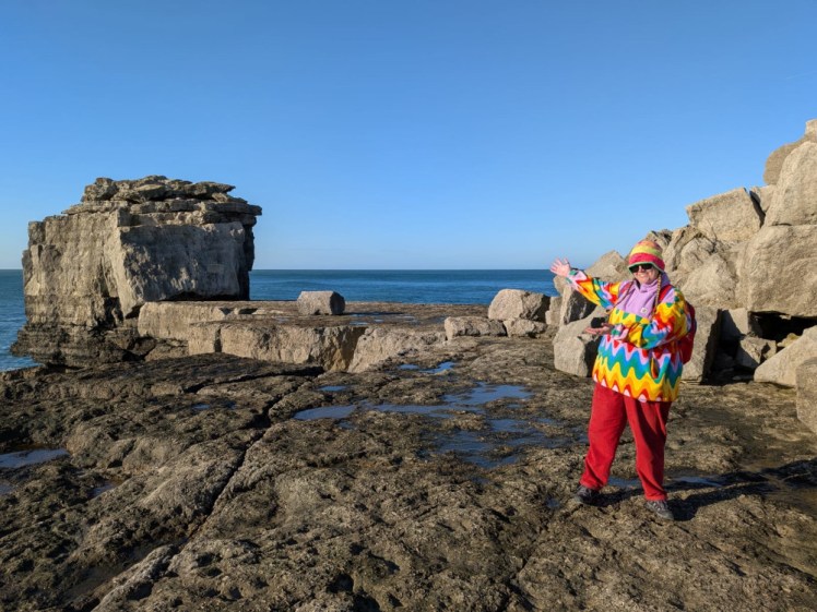 Me, in red trousers, rainbow fleece and rainbow hat, standing in front of a large rock apparently shaped like a Bible leaning on a pulpit.