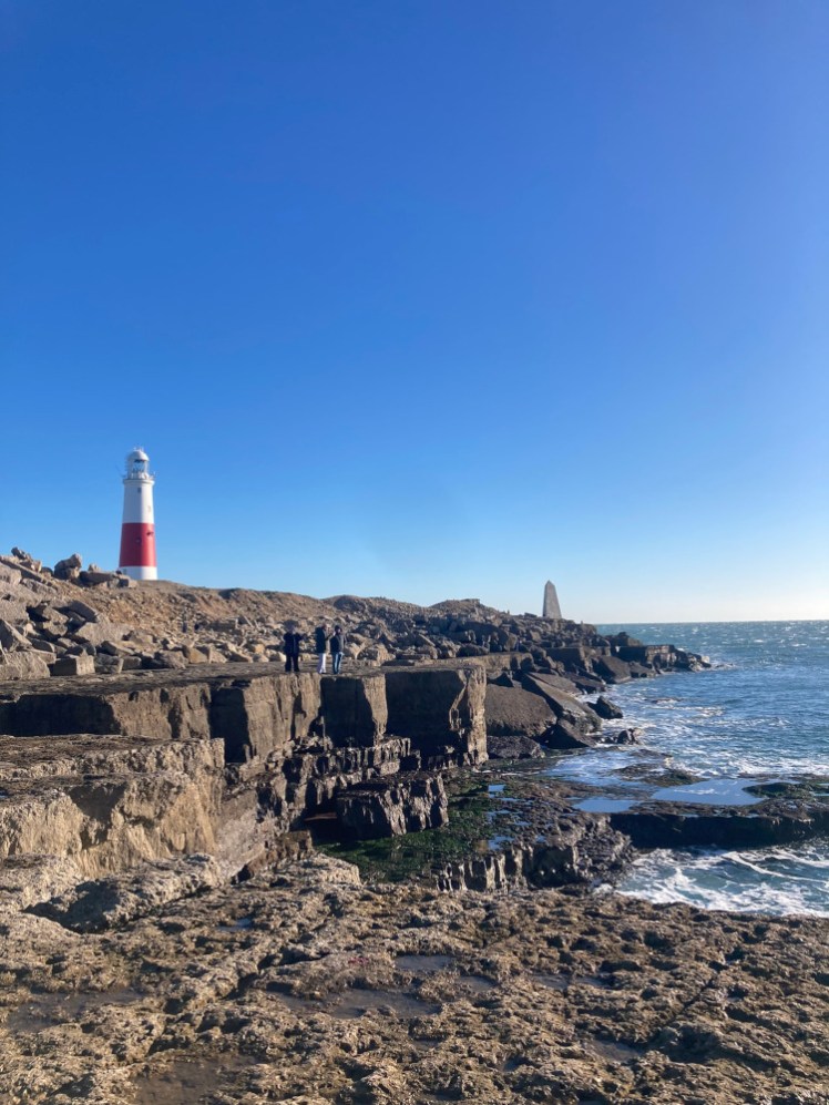 Portland Bill, looking along the low cliffs towards the lighthouse. The sea is calm but still splashing against the rock and the sky is blue.