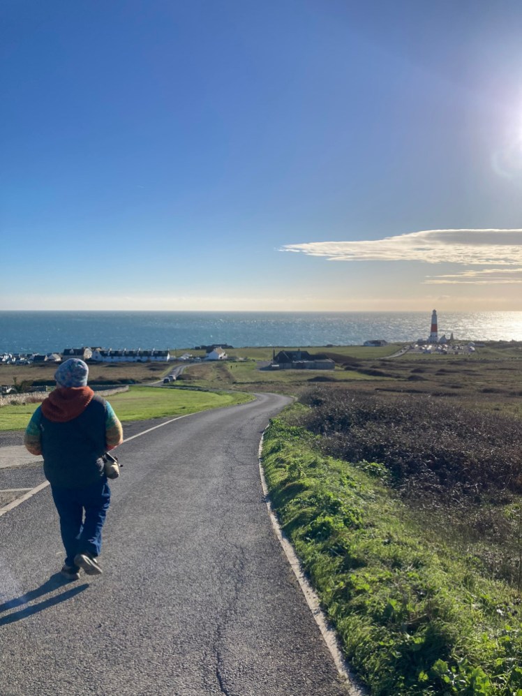 Tom walking down the road from the Old Higher Lighthouse with views down to the Bill and the open sea under a blue sky.