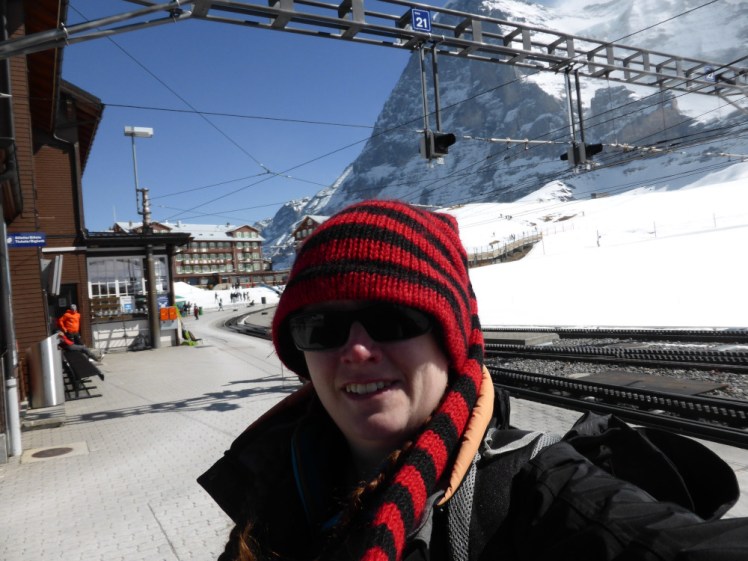 A selfie in the snow at Schynige Platte in early 2017. It's very snowy, there's a sheer mountain behind me and I'm wearing a red and black striped hat with a long tail.