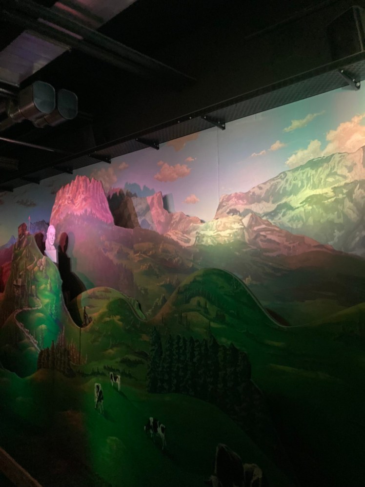 A 3D scene of Swiss hills and moutains made by several layers of scenery layered over each other with gaps in between to allow a jolly boy on a bike to cycle over the hills and the major chocolatiers of the 19th century to pop up.
