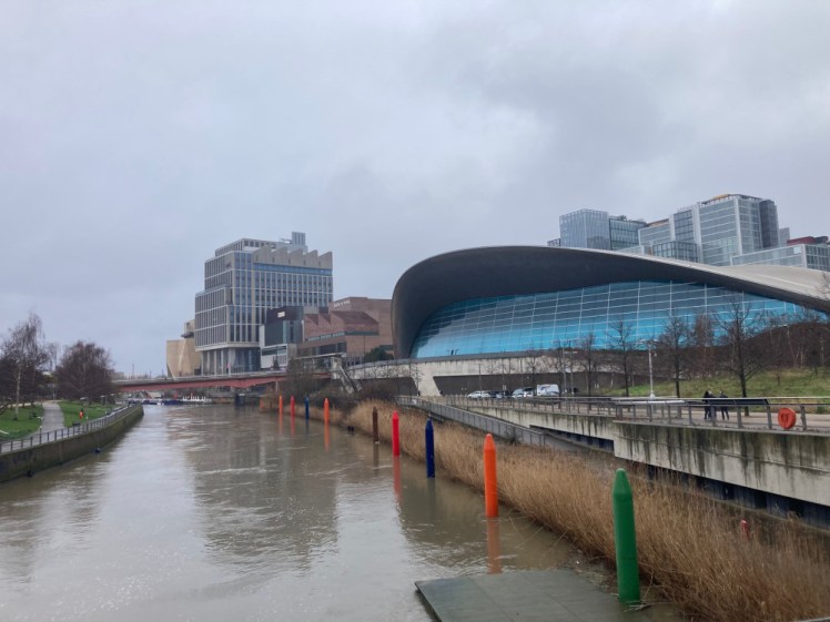 London Aquatics Centre, a building with a curved roof sticking out from the canal bank. A peaked roof shelters an entire array of bright blue glass panels that almost make it look like the mouth of a hungry fish from this angle.
