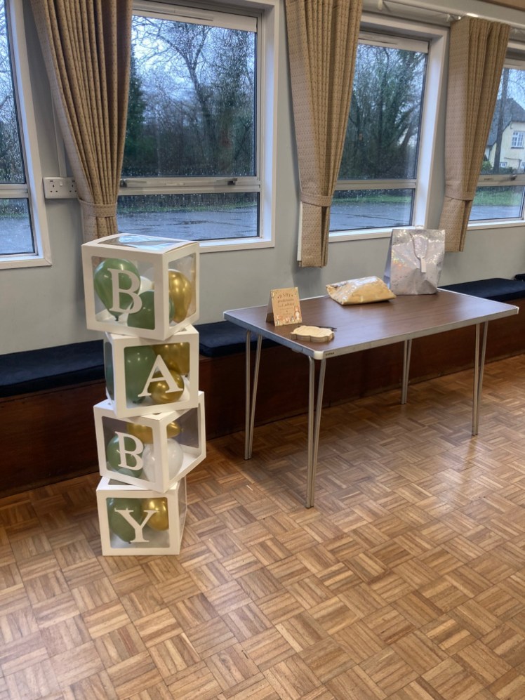 A table in a village hall. On it are two presents and a pile of baby prediction cards. Next to it are a pile of transparent boxes filled with balloons. The boxes have letters on the sides spelling out BABY from top to bottom.
