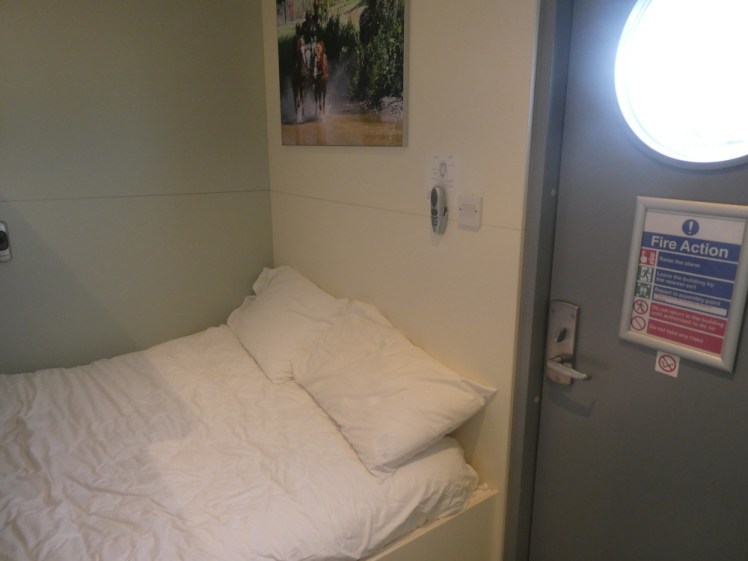 The main bed, which is a double with bedding, right next to the door which has a circular window in it.