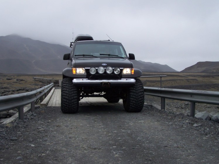 The enormous black jeep crossing a wooden bridge only a foot or two wider than it.