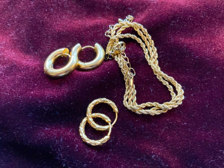 Two pairs of waterproof gold hoop earrings and a twisted chain necklace, all lying on a piece of purple velvet.