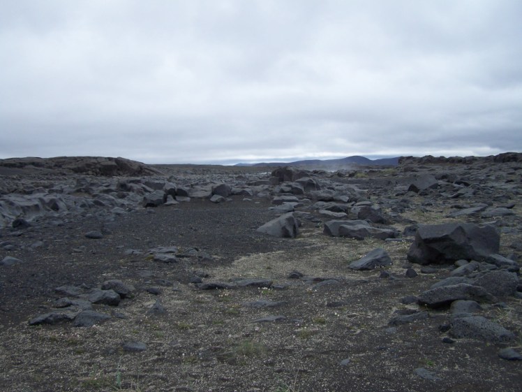 More lava field - lots of grey rock, a scattering of the very finest green grey vegetation amongst yellowish pumice powder and a bit of mist coming up from a minor waterfall hidden from this angle below the level of the lava field.