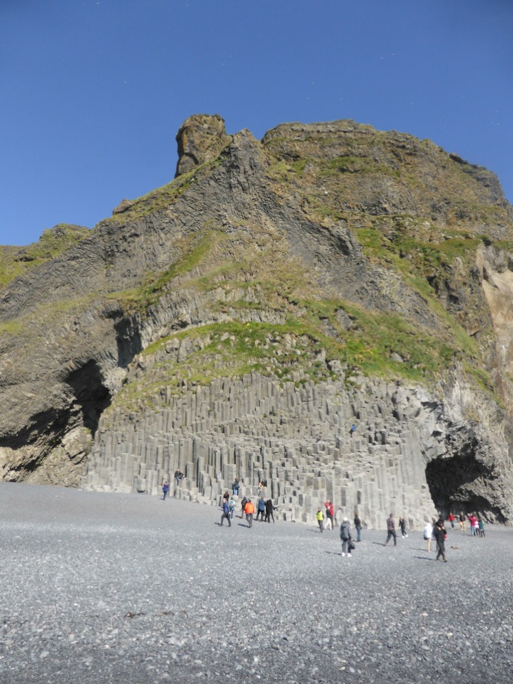The big green-covered basalt cliff at Reynisfjara, towering over tourists and black sand alike.